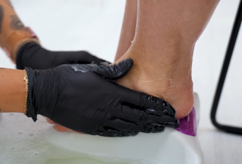 A professional wearing black gloves carefully scrubbing a person's heel during a pedicure treatment